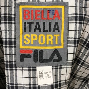 May include: Close-up of a vintage FILA jacket label. The label features the text "BIELLA ITALIA SPORT" in red, black, and yellow, with the FILA logo below. The background is a black and white plaid pattern.