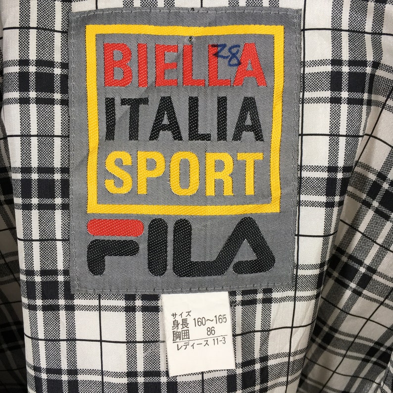 May include: Close-up of a vintage FILA jacket label. The label features the text "BIELLA ITALIA SPORT" in red, black, and yellow, with the FILA logo below. The background is a black and white plaid pattern.