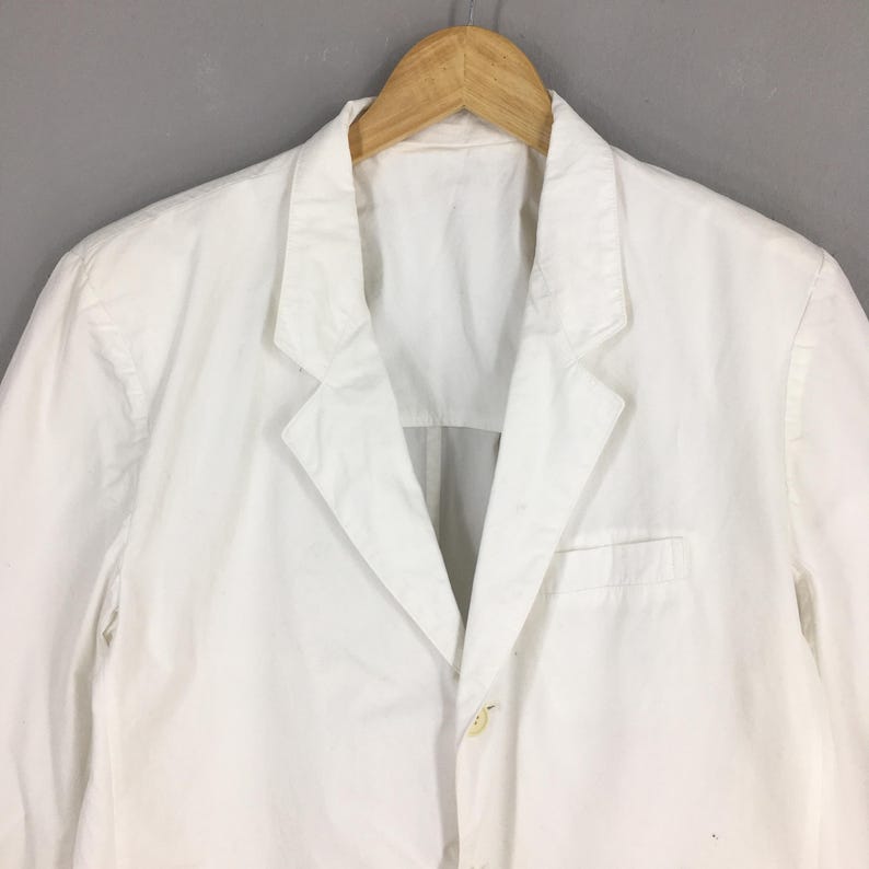 May include: A solid white blazer featuring a notched lapel, a single front button closure, and a chest pocket. The garment is displayed on a wooden hanger, showcasing a clean, structured design with long sleeves and a classic tailored silhouette.