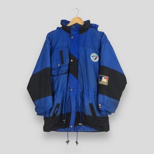 May include: A vintage blue and black jacket with a hood. The jacket features a Toronto Blue Jays logo patch and a Major League Baseball logo. It has a drawstring waist and pockets. The jacket is suspended from a wooden hanger. The jacket is a retro style.