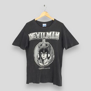 May include: Dark gray t-shirt featuring the word "DEVILMAN" in a white, distressed font above a circular graphic. The graphic depicts a character with a stern expression. The shirt is hanging on a wooden hanger.