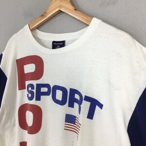 May include: White t-shirt with blue and red lettering that reads "POLO SPORT" with an American flag graphic. The shirt has blue and navy blue color blocking on the sleeves.