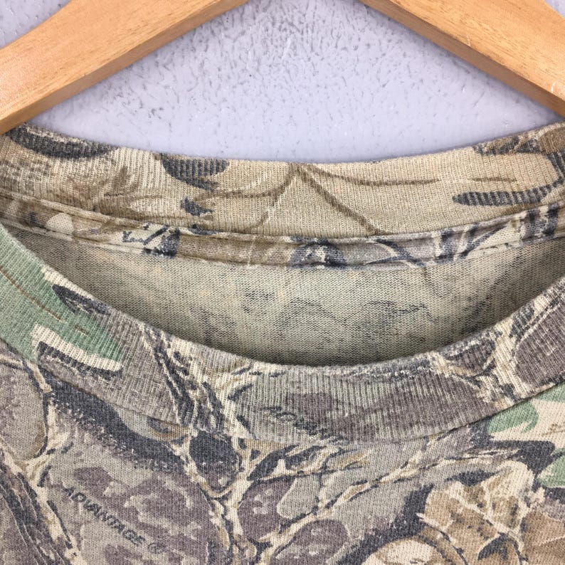 May include: Close-up of a camouflage pattern t-shirt. The shirt features a blend of green, brown, and gray hues, creating a woodland camouflage design. The text "ADVANTAGE" is visible at the bottom of the shirt.