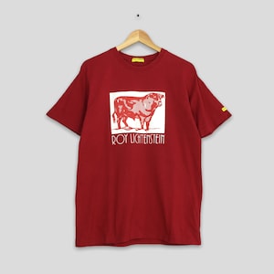 May include: A maroon t-shirt featuring a white square with a red illustration of a bull. Below the bull is the text "ROY LICHTENSTEIN" in white. The shirt is hanging on a wooden hanger.