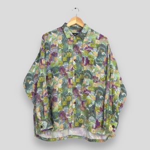 Puede incluir: Una camisa de manga larga con un colorido estampado abstracto en tonos verdes, morados, amarillos y grises. La camisa está colgada de una percha de madera. Tiene un bolsillo en el pecho izquierdo.