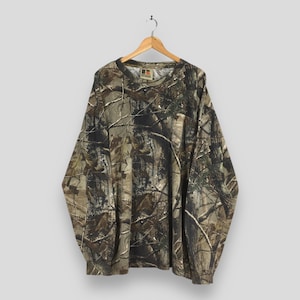 May include: Long-sleeved camouflage shirt in shades of brown, green, and tan. The shirt is hanging on a wooden hanger. The pattern mimics a forest environment.