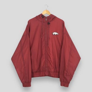 May include: A maroon zip-up windbreaker jacket featuring a hood with drawstring cords and a white embroidered razorback hog logo on the left chest. The jacket has long sleeves with elastic cuffs and a matching elastic hem at the bottom waist.