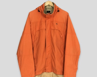 Pode incluir: Casaco laranja com capuz e interior bege, pendurado num cabide de madeira. O casaco tem fecho de correr, bolsos e um pequeno logótipo no peito. Um item de roupa exterior prático.