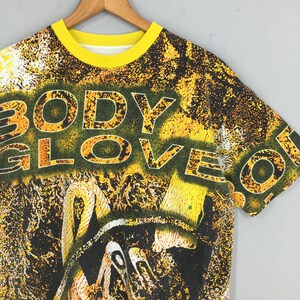 Peut inclure: Un t-shirt Body Glove jaune et noir avec un col jaune. Le t-shirt pr&eacute;sente les mots "BODY GLOVE" dans une police jaune et orange. Le graphisme est un m&eacute;lange de jaune, noir et blanc.