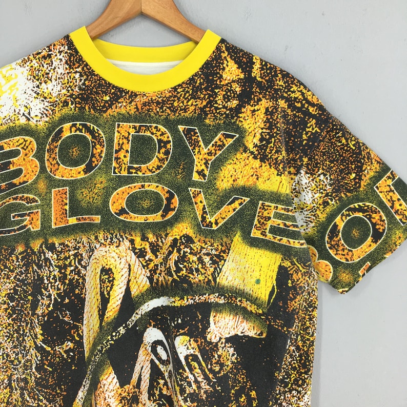 Peut inclure: Un t-shirt Body Glove jaune et noir avec un col jaune. Le t-shirt pr&eacute;sente les mots "BODY GLOVE" dans une police jaune et orange. Le graphisme est un m&eacute;lange de jaune, noir et blanc.