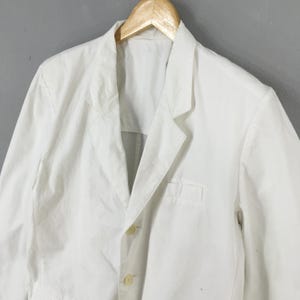 May include: A crisp white blazer featuring a notched lapel, a single breast pocket, and two light-colored buttons down the front. The garment is displayed on a wooden hanger, showcasing a structured collar and long sleeves with a clean, solid finish.