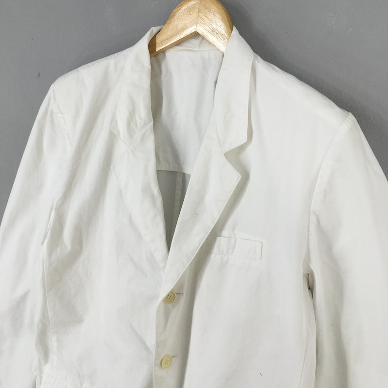 May include: A crisp white blazer featuring a notched lapel, a single breast pocket, and two light-colored buttons down the front. The garment is displayed on a wooden hanger, showcasing a structured collar and long sleeves with a clean, solid finish.