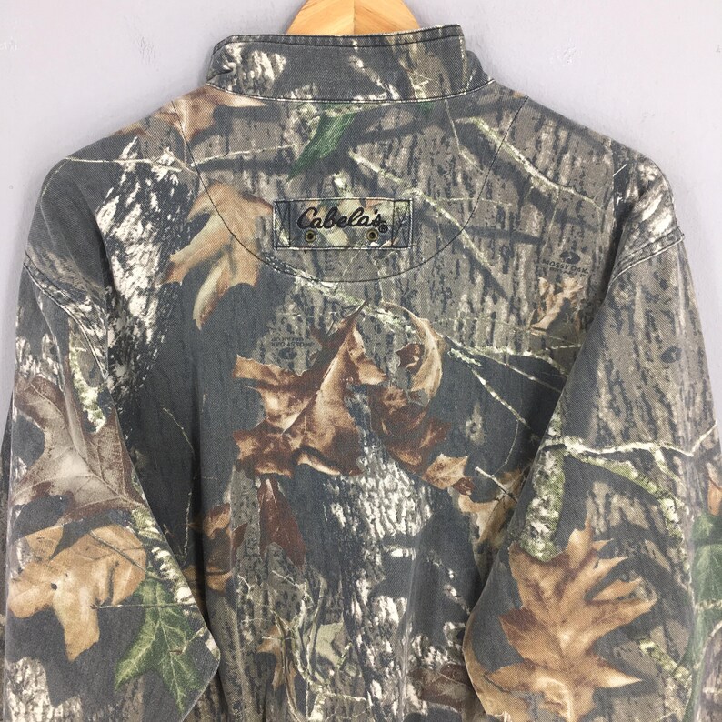 Op de afbeelding: Een langarmig shirt met camouflageprint en een Cabela's logo op de rug.