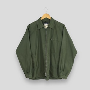 Puede incluir: Chaqueta verde oliva con cremallera, cuello y mangas largas, colgada de una percha de madera. La chaqueta tiene un pequeño bolsillo en el pecho y la marca "OLYMPIC" es visible en una etiqueta.