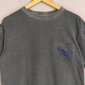 May include: A gray t-shirt with a pocket on the chest. The pocket has the blue text "Avirex U.S.A." The shirt is hanging on a wooden hanger.