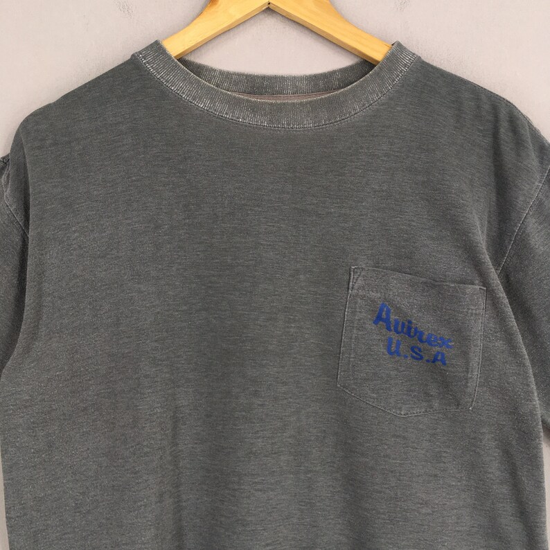 May include: A gray t-shirt with a pocket on the chest. The pocket has the blue text "Avirex U.S.A." The shirt is hanging on a wooden hanger.