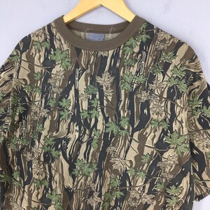 May include: A short-sleeved t-shirt with a camouflage pattern featuring brown, green, and black tones. The shirt has a brown collar and the text "Smoky Branch" is visible throughout the design. The shirt is hanging on a wooden hanger.