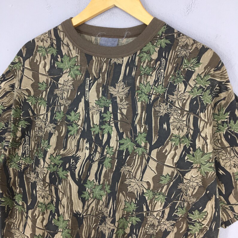 May include: A short-sleeved t-shirt with a camouflage pattern featuring brown, green, and black tones. The shirt has a brown collar and the text "Smoky Branch" is visible throughout the design. The shirt is hanging on a wooden hanger.