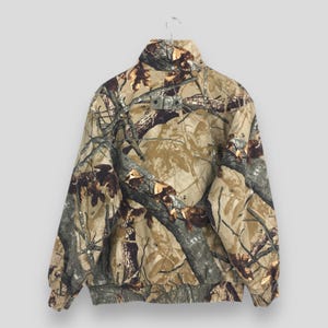 May include: A camouflage jacket with a high collar and a ribbed hem and cuffs. The jacket features a pattern of brown, tan, and green, resembling a forest environment. The jacket is hanging on a metal hanger.