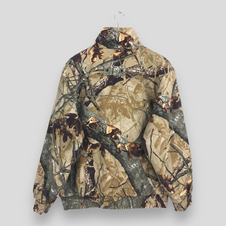May include: A camouflage jacket with a high collar and a ribbed hem and cuffs. The jacket features a pattern of brown, tan, and green, resembling a forest environment. The jacket is hanging on a metal hanger.