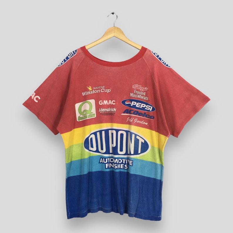 Op de afbeelding: Rood, geel, blauw en groen t-shirt met diverse logo's, waaronder "DUPONT AUTOMOTIVE FINISHES" en "PEPSI". Het shirt, met korte mouwen en een ronde hals, hangt aan een houten hanger.