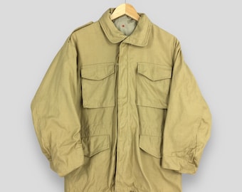 May include: A tan military-style jacket with four front pockets and a collared neckline. The jacket is hanging on a wooden hanger. The jacket has a drawstring at the bottom and epaulets on the shoulders.