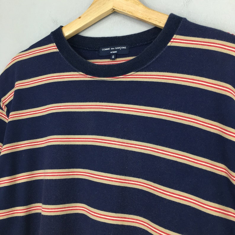 Puede incluir: Camiseta a rayas azul marino con rayas rojas y beige. La camiseta tiene cuello redondo y mangas cortas. La marca "COMME des GARCONS" es visible en la etiqueta.