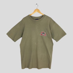 Op de afbeelding: Een olijfgroen t-shirt met korte mouwen, een ronde hals en een kleine afbeelding op de linkerborst. De afbeelding toont een zwart cartoon-kattengezicht met grote witte ogen en een brede grijns boven de rode tekst 26 RED.