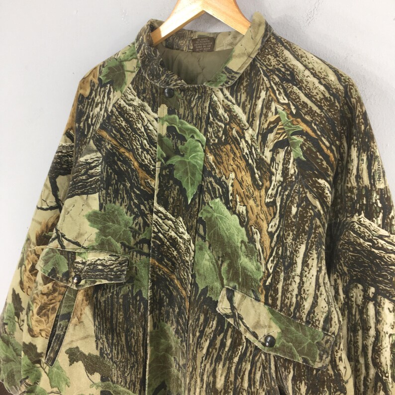 May include: Camouflage jacket with a button-up front, two front pockets, and a collar. The jacket features a pattern of brown, green, and tan, resembling a forest. The jacket is hanging on a wooden hanger.