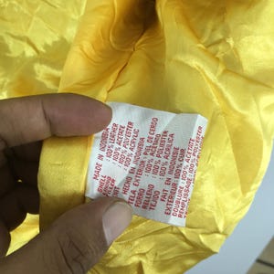 May include: A close-up shot of the inside of a yellow garment, showing a label detailing the materials: 100% leather shell, 100% acetate lining, and 100% polyester filler. The label states "Made in Indonesia."