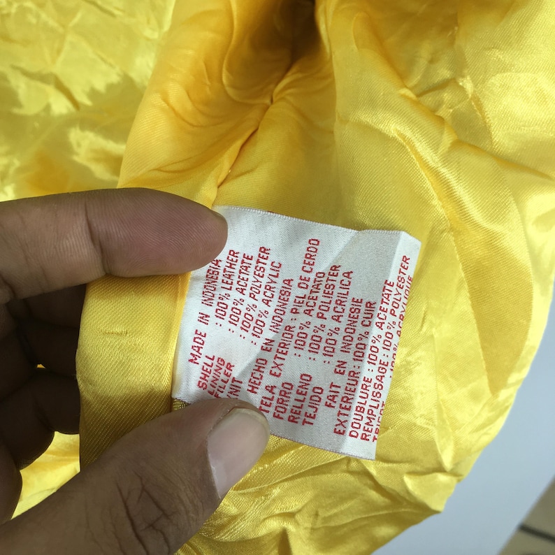 May include: A close-up shot of the inside of a yellow garment, showing a label detailing the materials: 100% leather shell, 100% acetate lining, and 100% polyester filler. The label states "Made in Indonesia."
