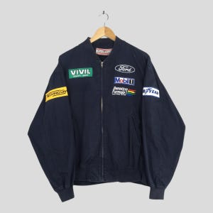Puede incluir: Chaqueta bomber azul marino con múltiples parches de patrocinadores, incluyendo "Vivil", "Ford" y "Mobil 1". La chaqueta tiene cremallera completa y puños y dobladillo acanalados. Cuelga de una percha de madera.