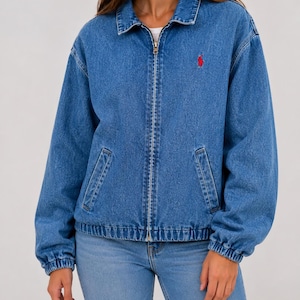 May include: A medium-wash blue denim jacket featuring a classic collar, a full-length metal front zipper, and long sleeves with elasticized cuffs. A small red embroidered logo is positioned on the upper left chest of the garment.