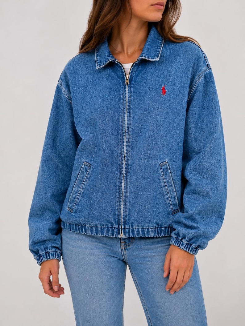 May include: A medium-wash blue denim jacket featuring a classic collar, a full-length metal front zipper, and long sleeves with elasticized cuffs. A small red embroidered logo is positioned on the upper left chest of the garment.