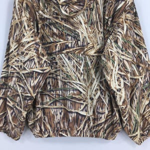 Puede incluir: Chaqueta de camuflaje con un patr&oacute;n de hierba alta en tonos marrones, beige y verdes. La chaqueta tiene cintura ajustable con cord&oacute;n, cierre de cremallera y botones. La tela parece ligera.