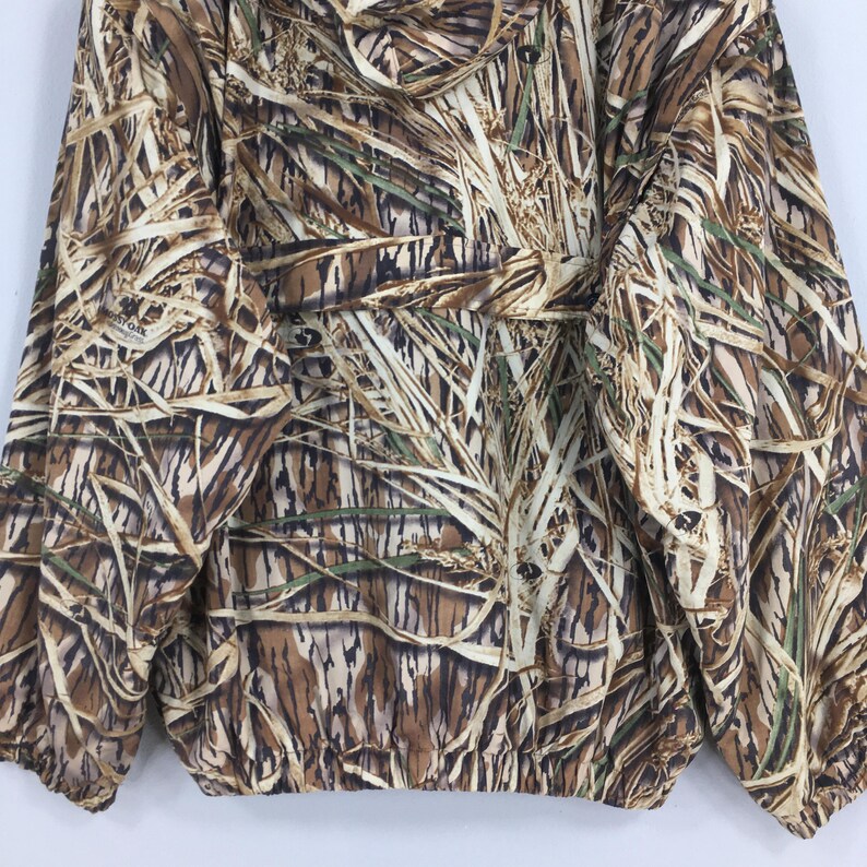 Puede incluir: Chaqueta de camuflaje con un patr&oacute;n de hierba alta en tonos marrones, beige y verdes. La chaqueta tiene cintura ajustable con cord&oacute;n, cierre de cremallera y botones. La tela parece ligera.