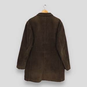 Puede incluir: Un abrigo de pana marr&oacute;n colgado de una percha de madera. El abrigo tiene cierre de botones y cuello cl&aacute;sico. La tela tiene una textura acanalada. El abrigo es de color marr&oacute;n liso.