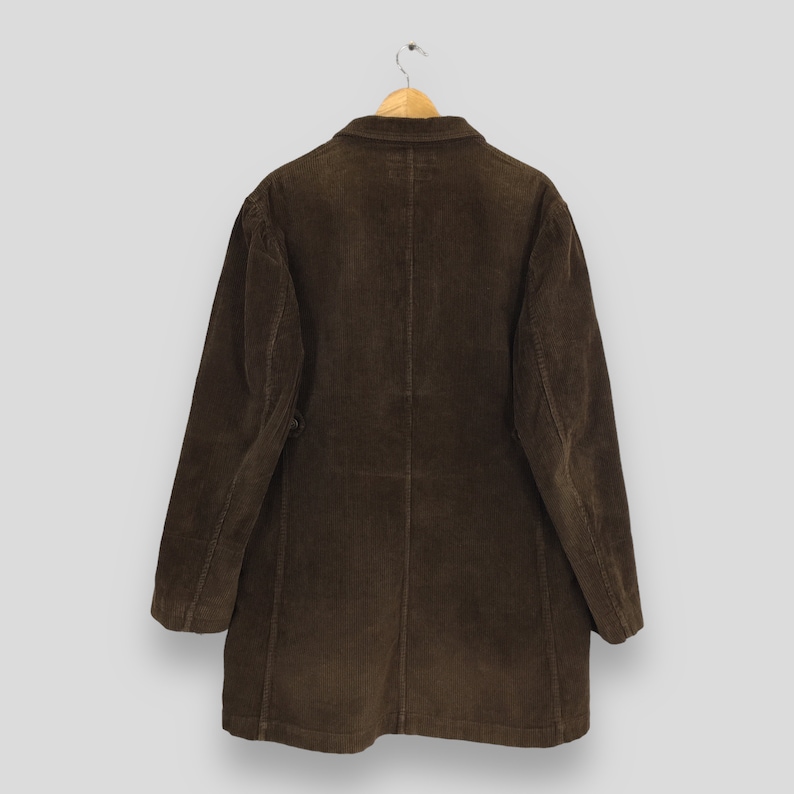 Puede incluir: Un abrigo de pana marr&oacute;n colgado de una percha de madera. El abrigo tiene cierre de botones y cuello cl&aacute;sico. La tela tiene una textura acanalada. El abrigo es de color marr&oacute;n liso.