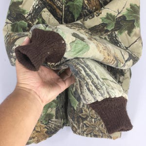 May include: Camouflage jacket with brown ribbed cuffs. The jacket features a pattern of green, brown, and tan, resembling a forest environment. The sleeve is folded, revealing the interior lining. The Realtree logo is visible on the sleeve.