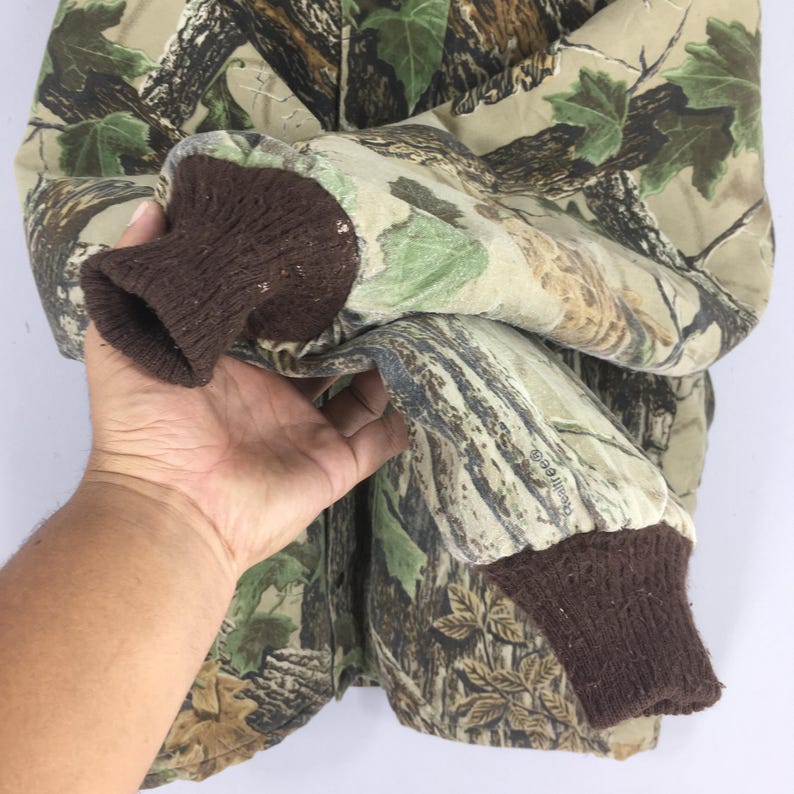 May include: Camouflage jacket with brown ribbed cuffs. The jacket features a pattern of green, brown, and tan, resembling a forest environment. The sleeve is folded, revealing the interior lining. The Realtree logo is visible on the sleeve.