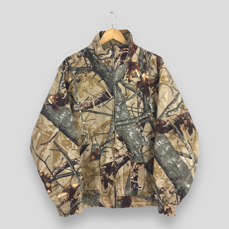 May include: A full-zip camouflage jacket with a high collar, elastic cuffs, and a waistband. The jacket features a pattern of brown, tan, and gray tree branches and leaves. It is hanging on a wooden hanger.