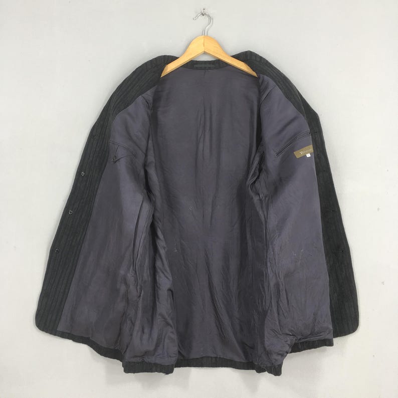 May include: A dark charcoal gray blazer featuring a subtle vertical pinstripe pattern, a notched lapel, and a solid dark gray interior lining. The garment is displayed open on a wooden hanger, showcasing the internal structure and button closure.