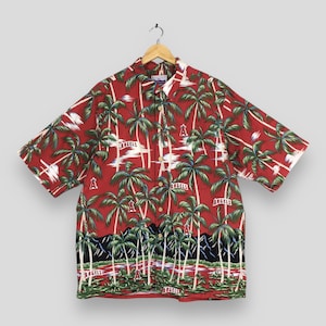 May include: Red short-sleeve button-down shirt with a tropical palm tree pattern. The shirt features the word "ANGELS" and the team logo. The shirt is hanging on a wooden hanger.