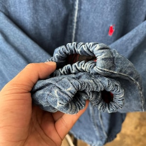 May include: A blue denim jacket featuring elasticized cuffs with gathered fabric. The jacket has a red embroidered logo on the chest and a visible red plaid lining inside the sleeve cuffs. The denim material shows a medium blue wash finish.
