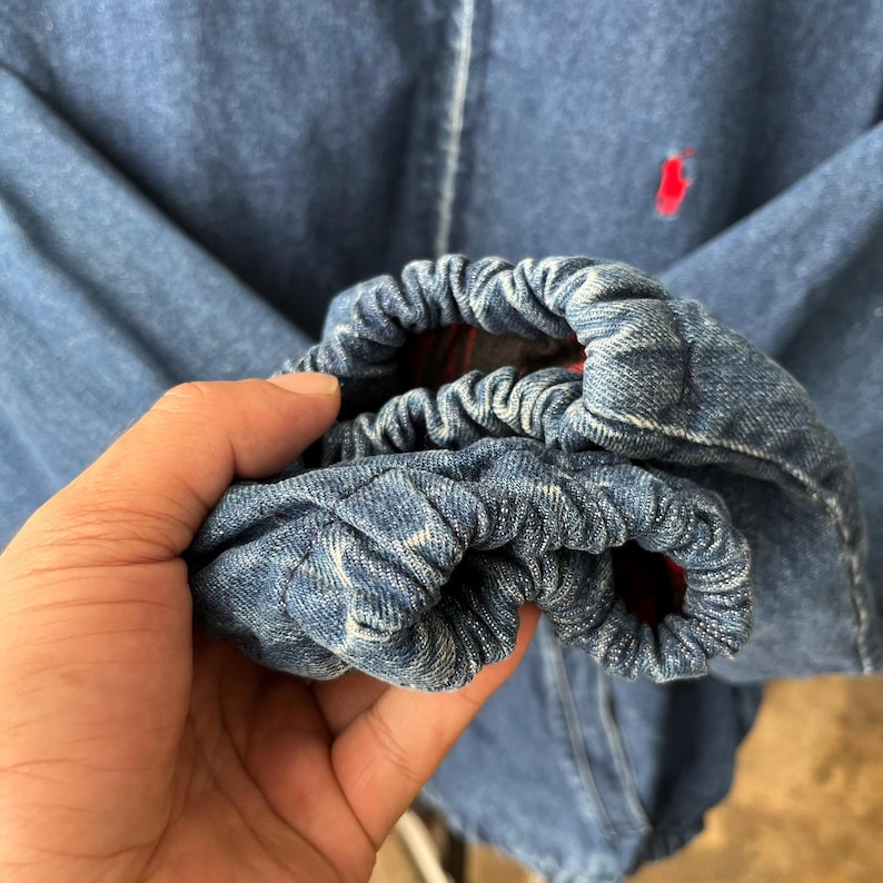 May include: A blue denim jacket featuring elasticized cuffs with gathered fabric. The jacket has a red embroidered logo on the chest and a visible red plaid lining inside the sleeve cuffs. The denim material shows a medium blue wash finish.