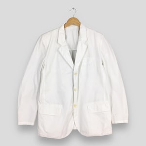May include: A solid white blazer featuring a notched lapel, long sleeves, and a three-button front closure. The garment includes two rectangular flap pockets positioned at the lower front and is displayed on a wooden hanger against a neutral background.