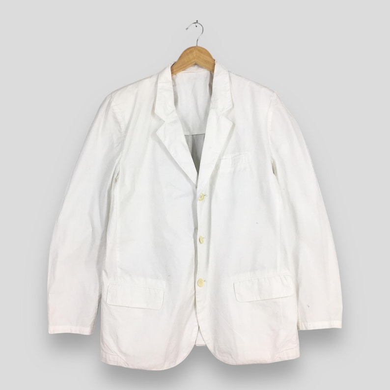 May include: A solid white blazer featuring a notched lapel, long sleeves, and a three-button front closure. The garment includes two rectangular flap pockets positioned at the lower front and is displayed on a wooden hanger against a neutral background.