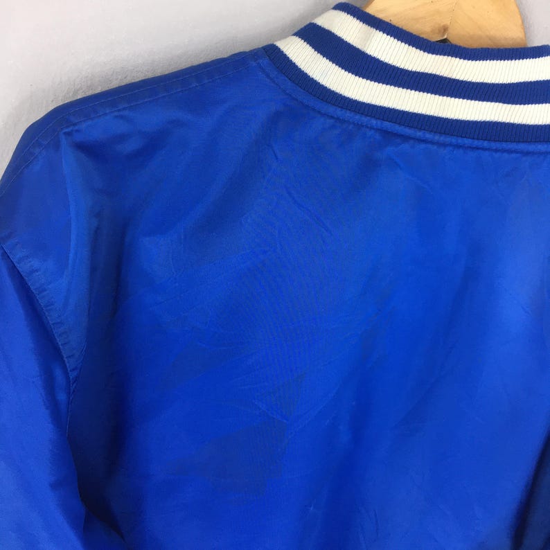 May include: A bright blue jacket featuring a ribbed collar with white stripes. The garment is constructed from a smooth, lightweight material and includes a high neckline that sits against the collarbone, showcasing a clean, solid-coloured design.