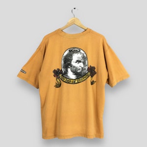 May include: A mustard yellow t-shirt with a graphic of a man's head inside a glass sphere. The graphic is surrounded by a banner that reads "Surfers of Fortune".
