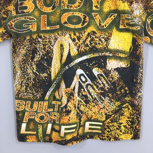 Peut inclure: T-shirt vintage avec un motif graphique avec les mots "BODY GLOVE" en jaune et vert. Le t-shirt porte &eacute;galement les mots "BUILT FOR LIFE" en jaune. Le motif comprend une corde et d'autres &eacute;l&eacute;ments abstraits.